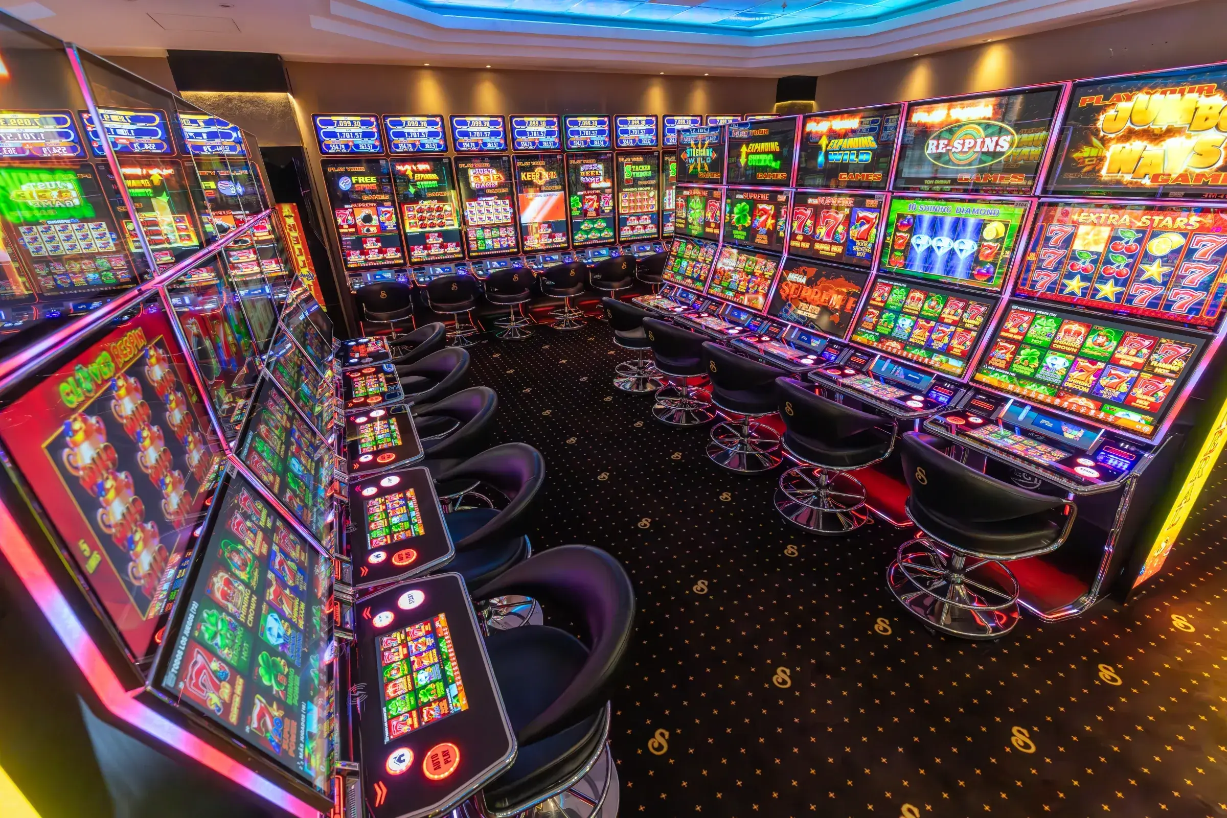 A Senator gaming room in Guatemala, machines lit around three walls with linked jackpot totals on the screens above one bank and monogrammed stools along the carpet