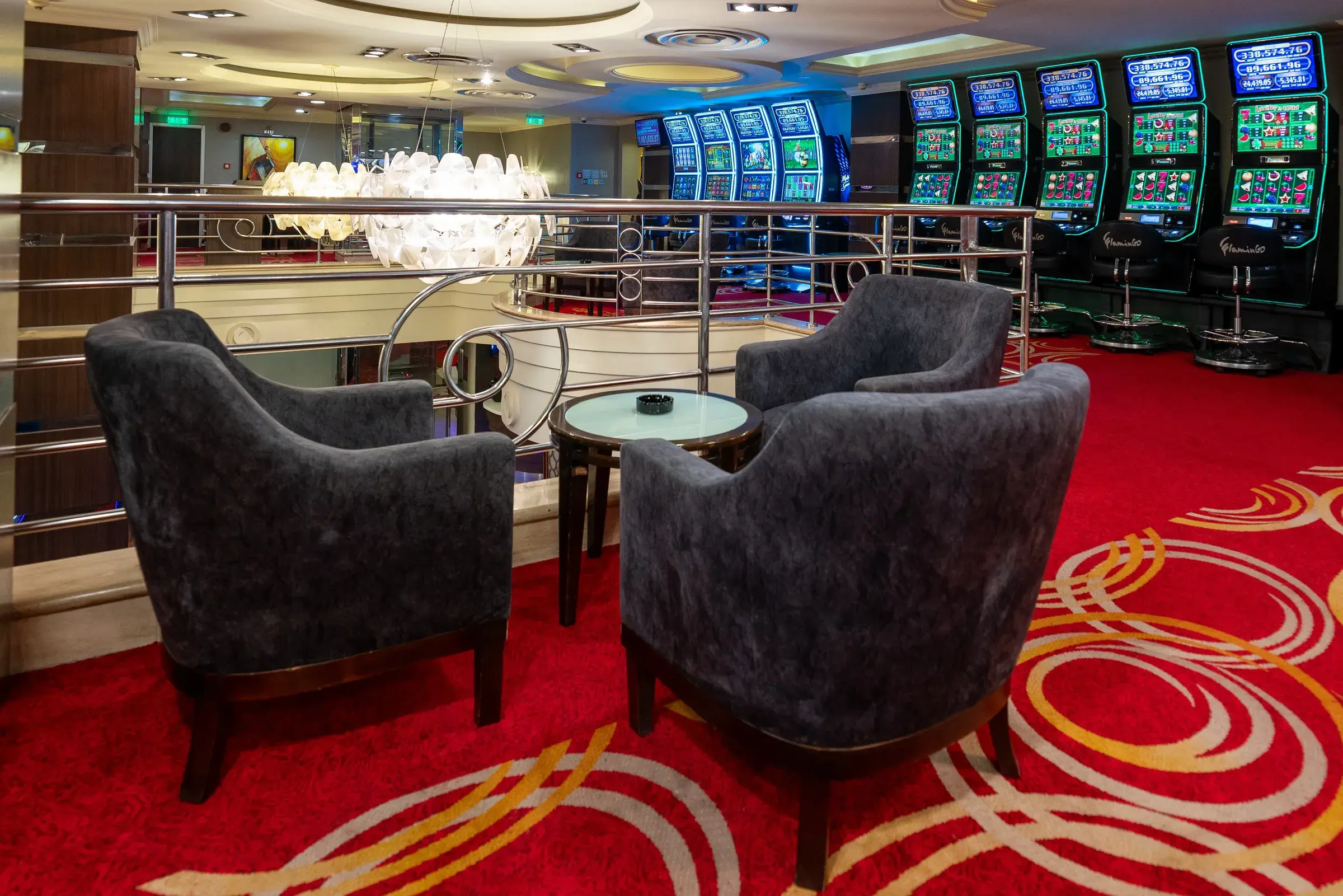 Four grey armchairs around a low table on the mezzanine behind a chrome rail, a chandelier at the left and a green lit slot bank beyond