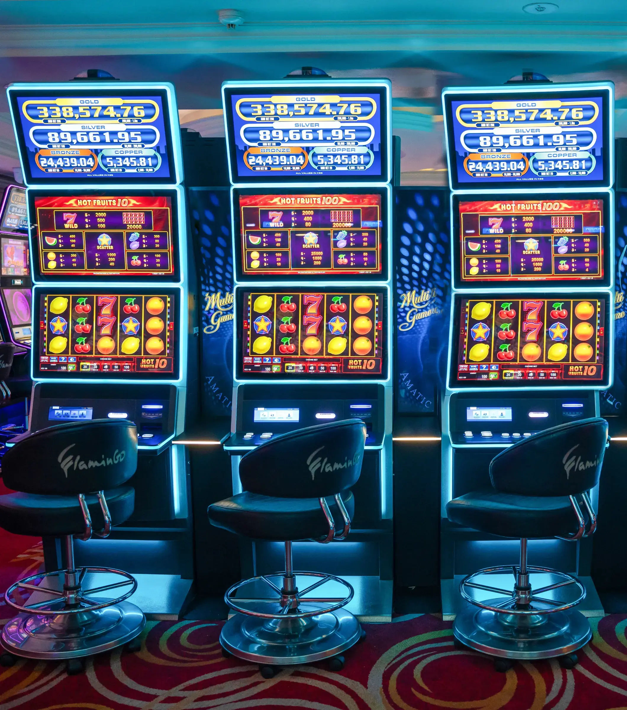 Three Hot Fruits terminals in blue neon with jackpot boards above them and Flamingo stools drawn up in front