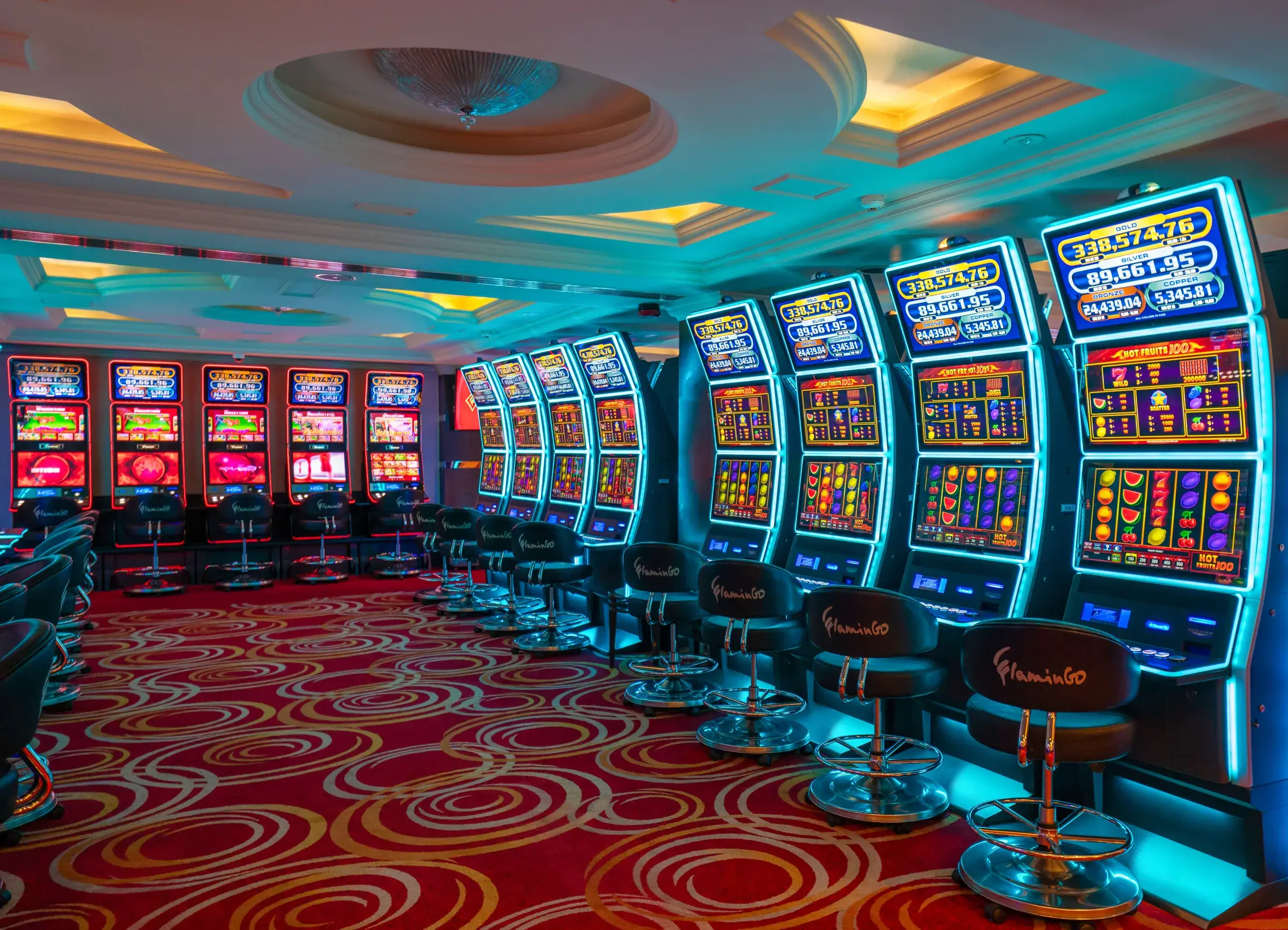 A corner of the slot floor where a red lit wall of machines meets a blue lit one, under the coffered ceiling