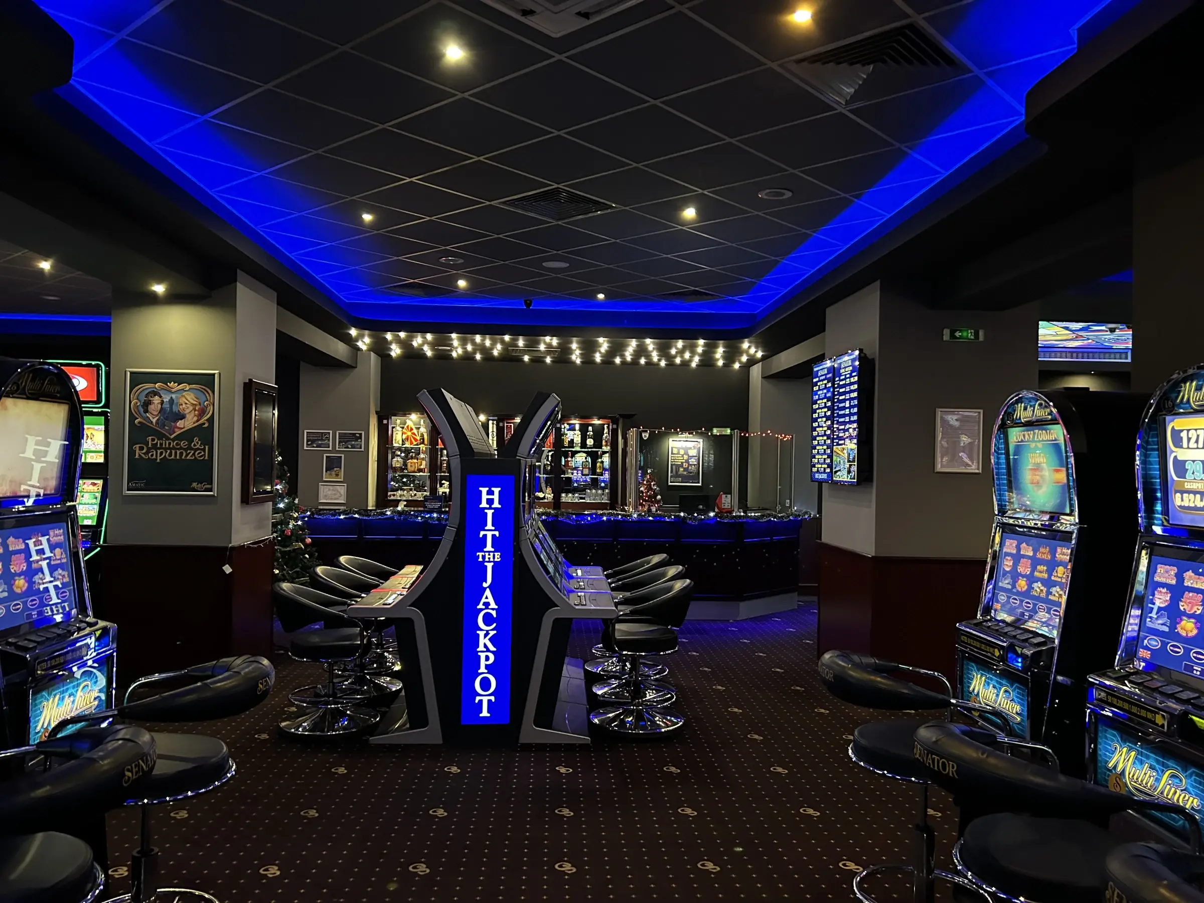 A lit Hit the Jackpot totem in the middle of a Senator club floor, the bar behind it and slot cabinets on both sides