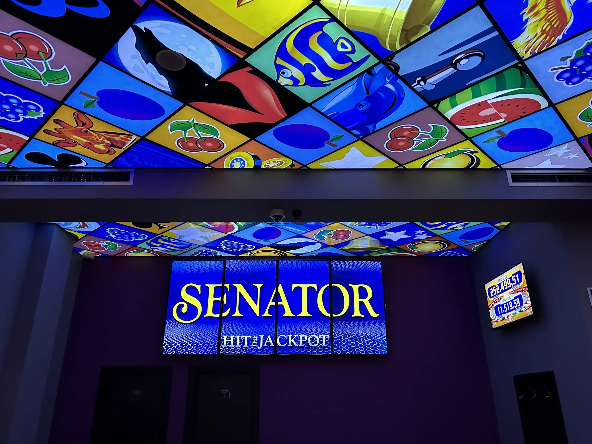 A backlit stained glass ceiling of fruit and card symbols above an illuminated Senator Hit the Jackpot sign