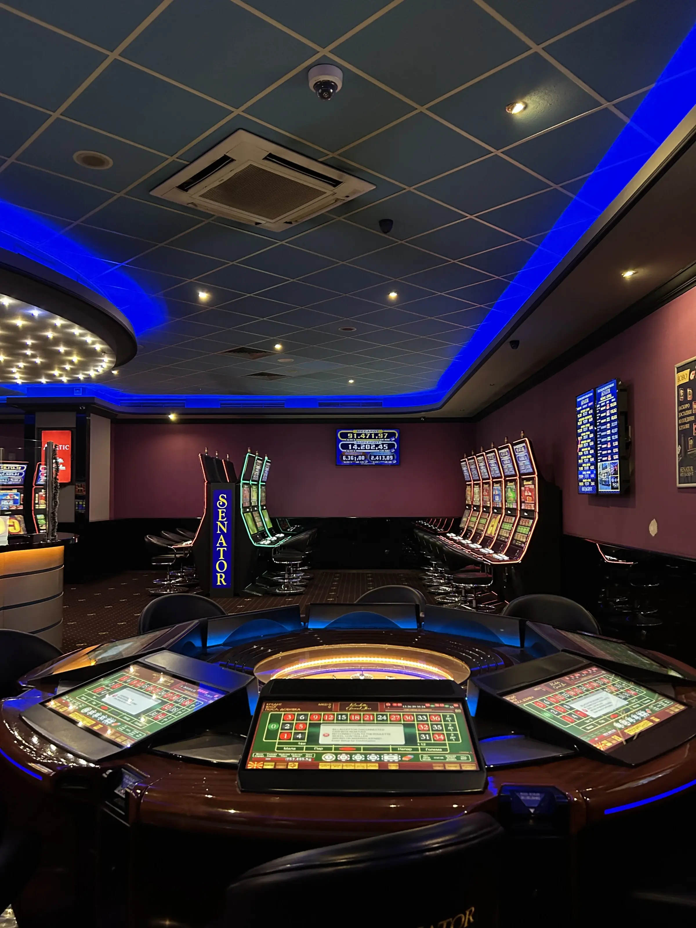 An electronic roulette table in a room lit by blue cove lighting, a Senator cabinet and a row of slot machines along the wall