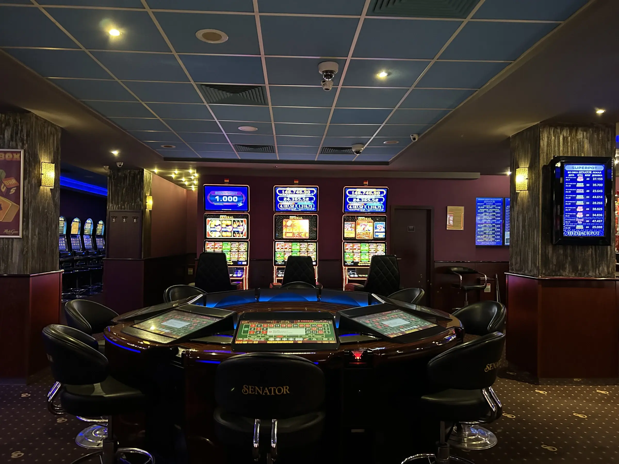 An electronic roulette table ringed by Senator branded chairs, with three slot cabinets and a jackpot board behind it