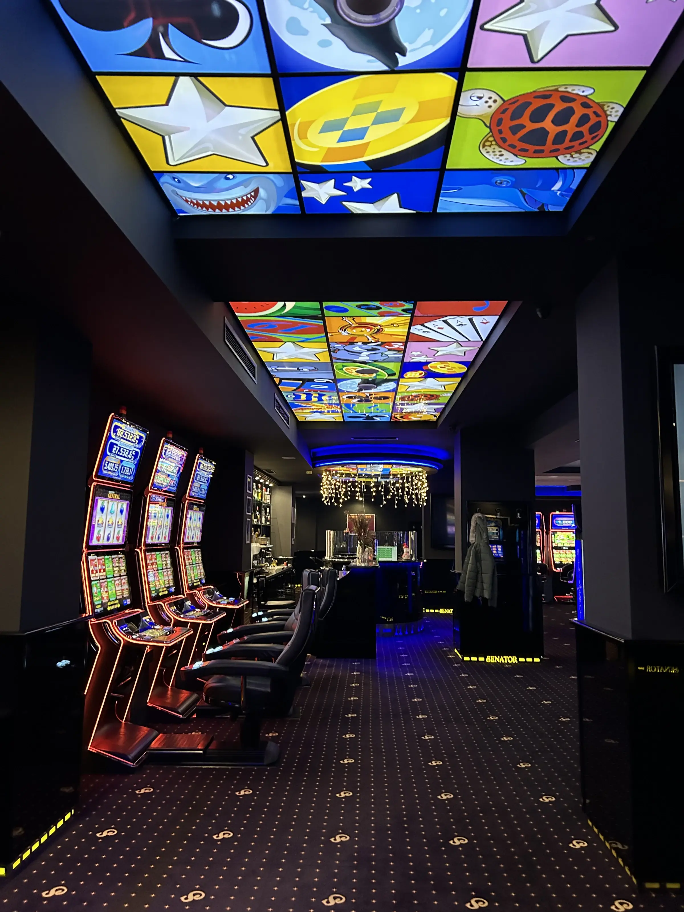 A corridor of slot machines under backlit printed glass ceiling panels, with a chandelier and the bar at the far end