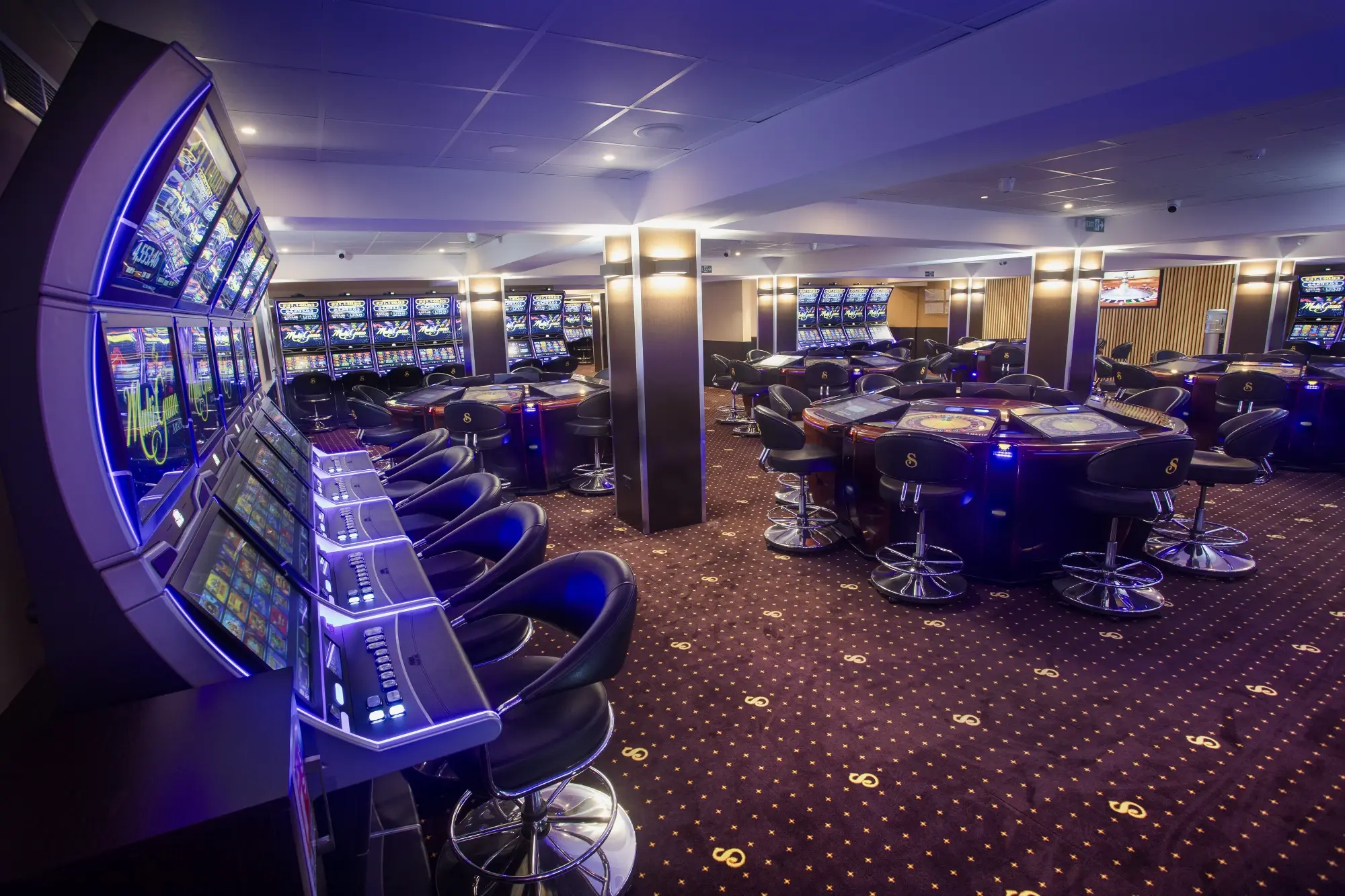 A curved line of slot cabinets and their stools in the foreground, with electronic roulette terminals further into the room, Senator Club, Mauritius